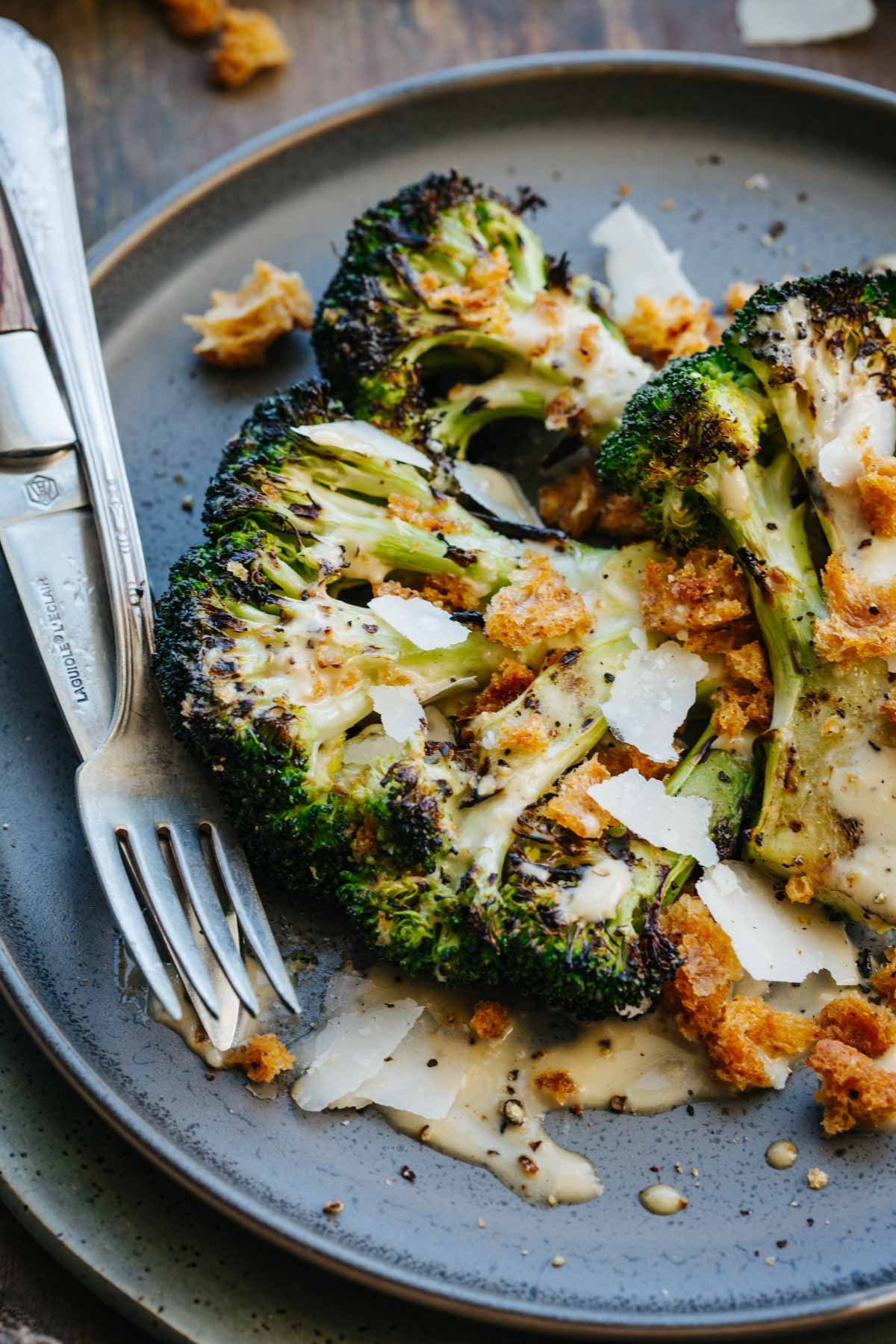Close-up of grilled broccoli florets on a plate, glossy with Caesar dressing and scattered with croutons, shaved Parmigiano, and black pepper, fork resting to the side.