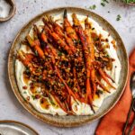 Over heat shot of plated honey glazed carrots, whipped ricotta and hazelnuts in a ceramic plate next to serving plates, cloth napkins, thyme and honey.