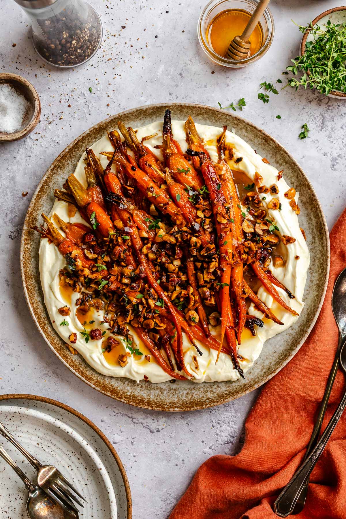 Overhead shot of plated honey glazed carrots, whipped ricotta and hazelnuts in a ceramic plate next to serving plates, cloth napkins, thyme and honey.