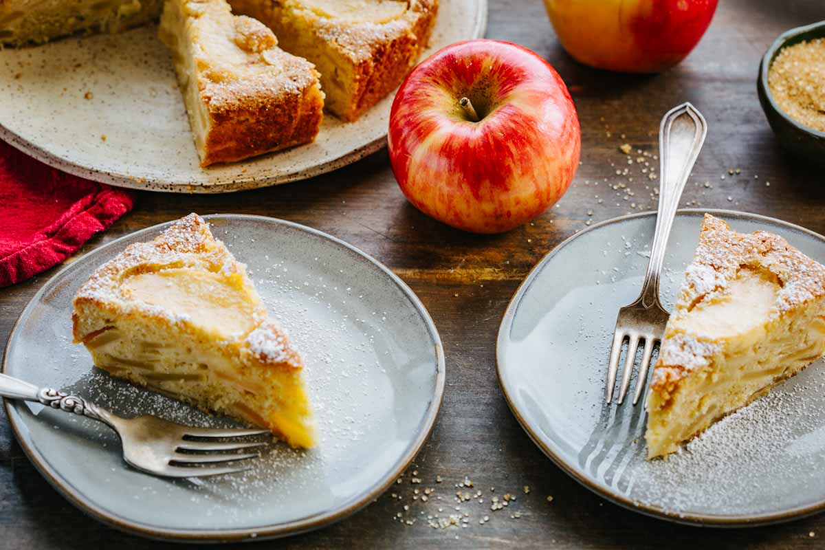 Two slices of Italian apple cake on gray plates with forks, next to a whole cake and fresh apples on a dark wood table.