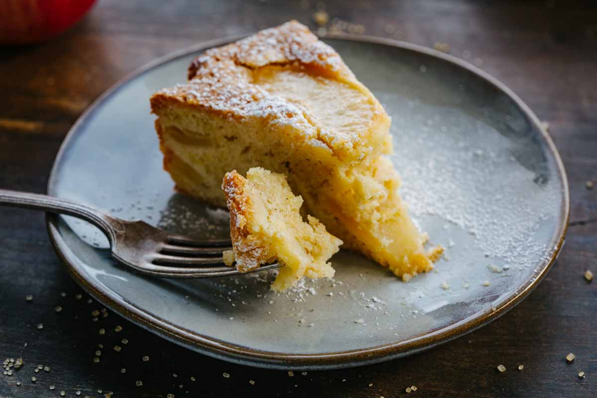 A slice of Italian apple cake on a gray plate with a bite on a fork, showing the tender crumb and apple layers inside.