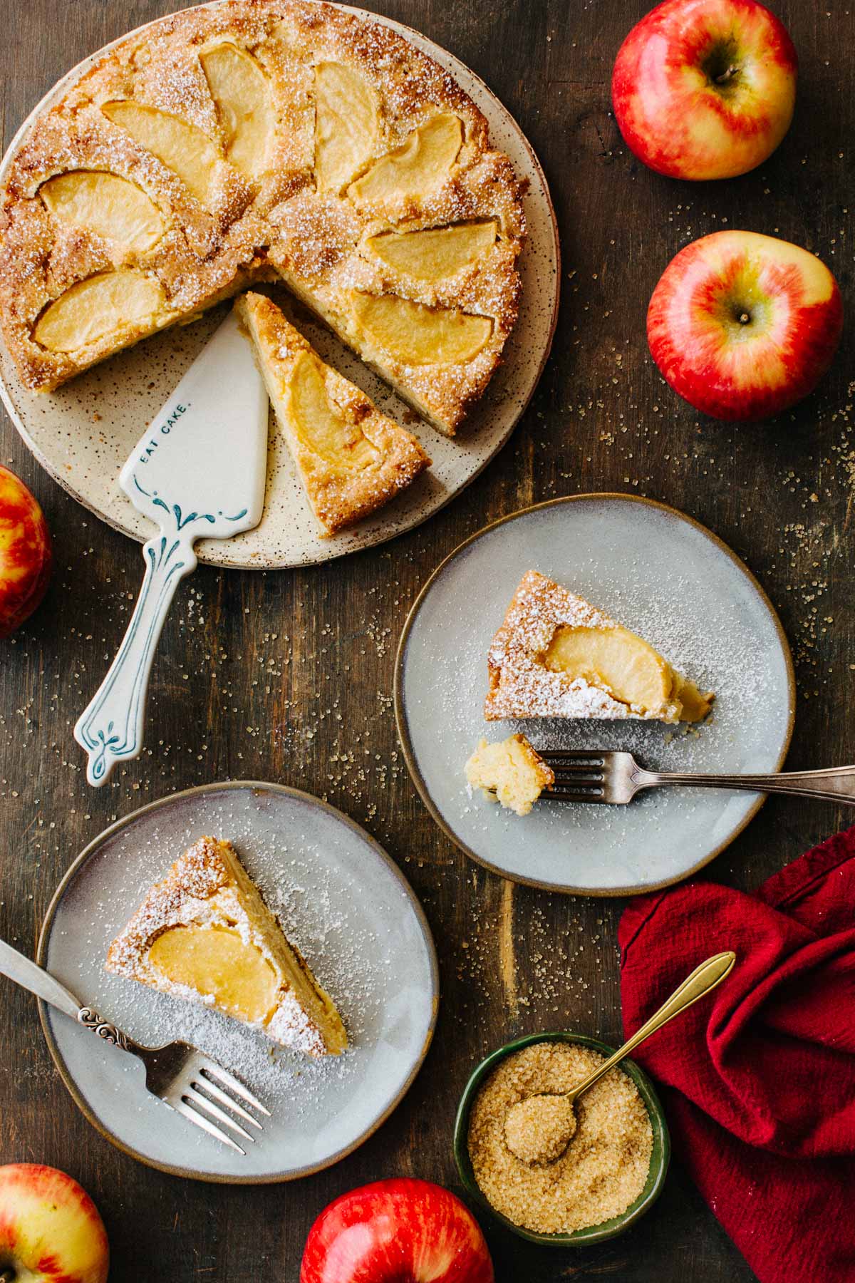 Overhead view of a sliced Italian apple cake with a cake server, plated slices, apples, and a bowl of raw sugar on a wood table.