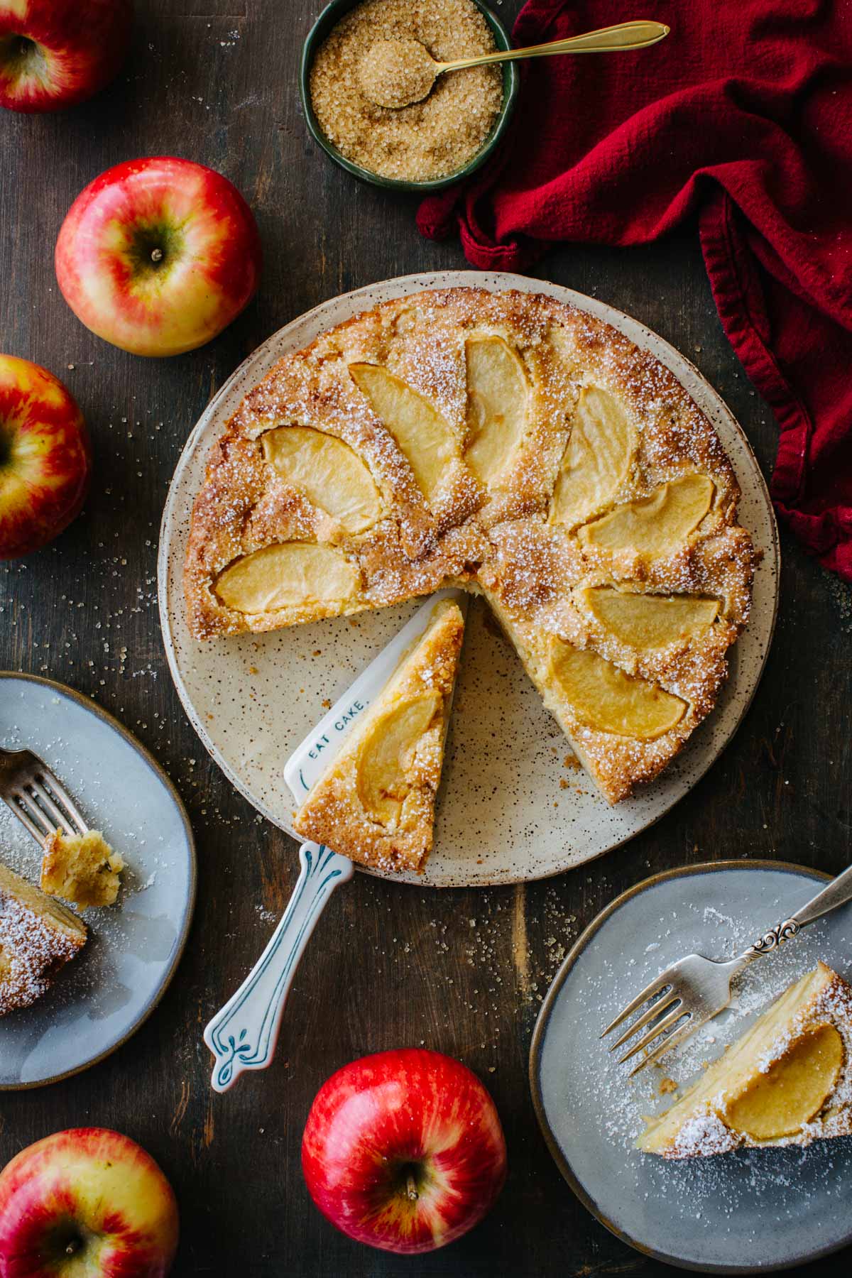 Overhead shot of a whole Italian apple cake with one slice cut and lifted, surrounded by apples, a red napkin, and a bowl of raw sugar.
