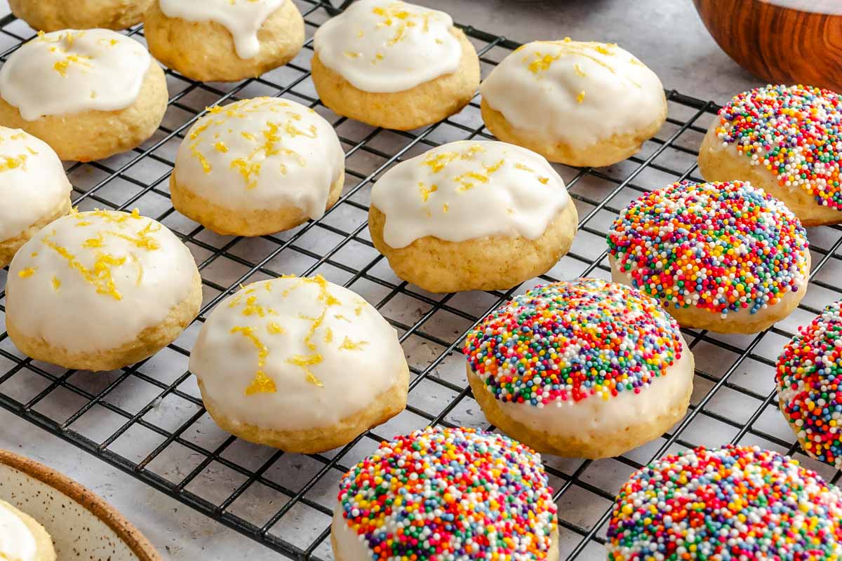 Lemon ricotta cookies cooling on a wire rack, some topped with lemon glaze and fresh lemon zest, others coated in rainbow nonpareil sprinkles.