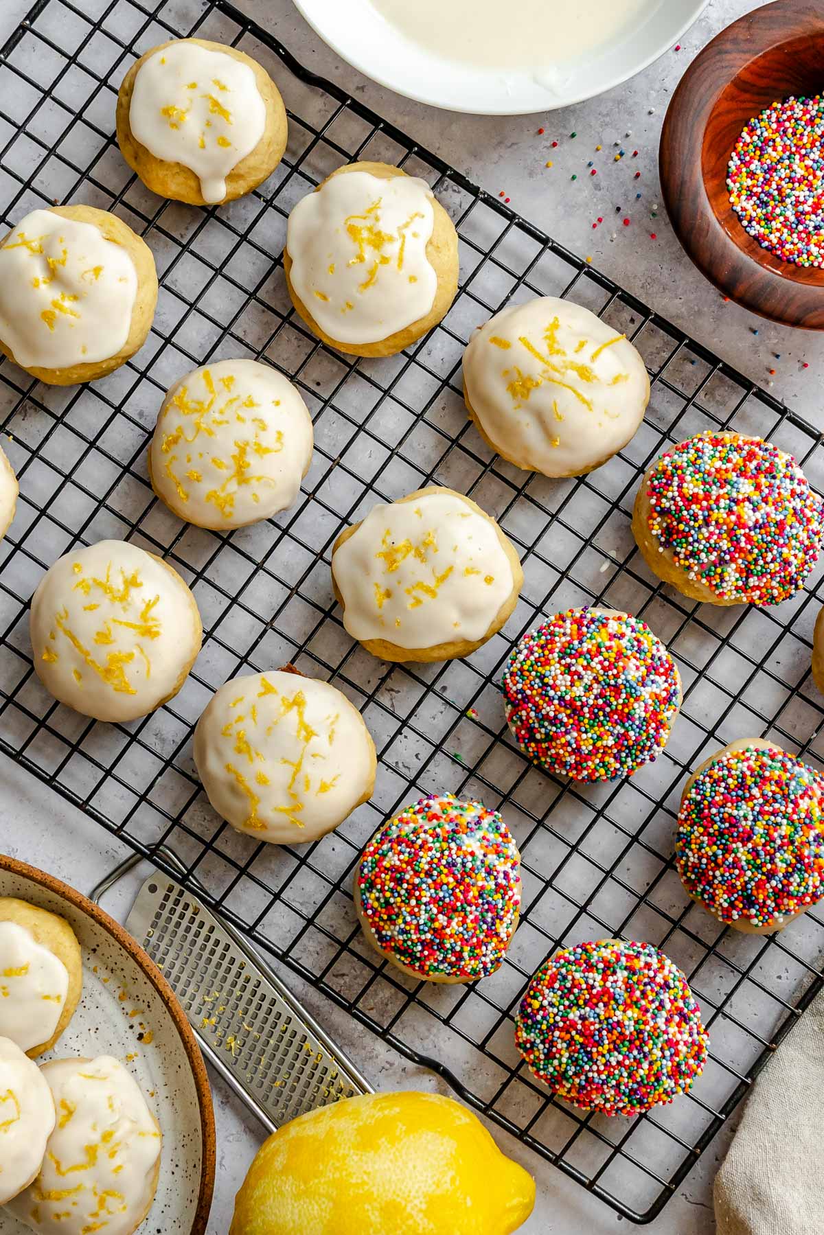 Close-up of glazed lemon ricotta cookies on a cooling rack, topped with lemon zest, next to a bowl of sprinkles and a whole lemon.