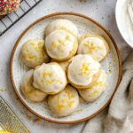 A plate stacked with lemon-glazed ricotta cookies topped with lemon zest, with a zester and fresh lemon in the foreground.