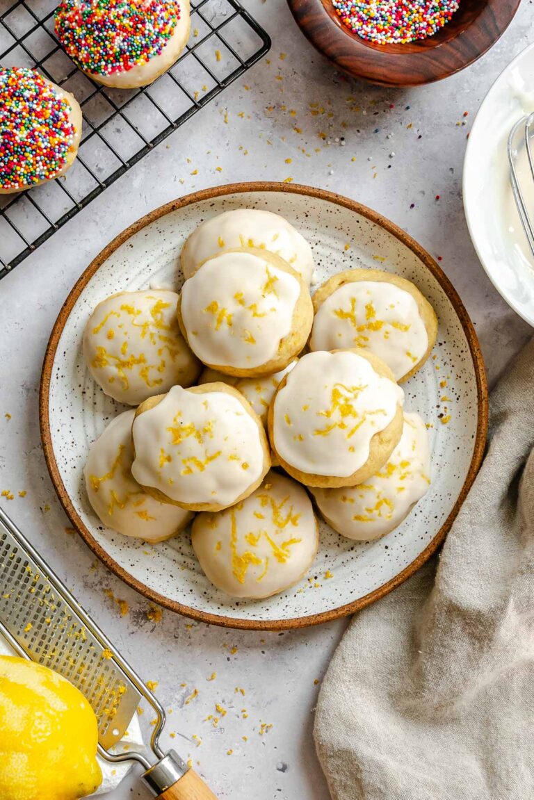 A plate stacked with lemon-glazed ricotta cookies topped with lemon zest, with a zester and fresh lemon in the foreground.