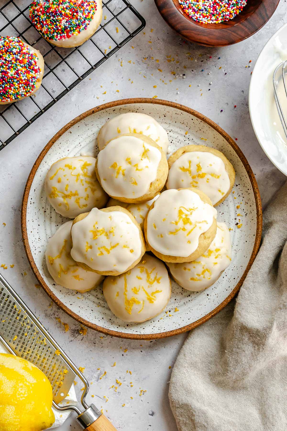 A plate stacked with lemon-glazed ricotta cookies topped with lemon zest, with a zester and fresh lemon in the foreground.