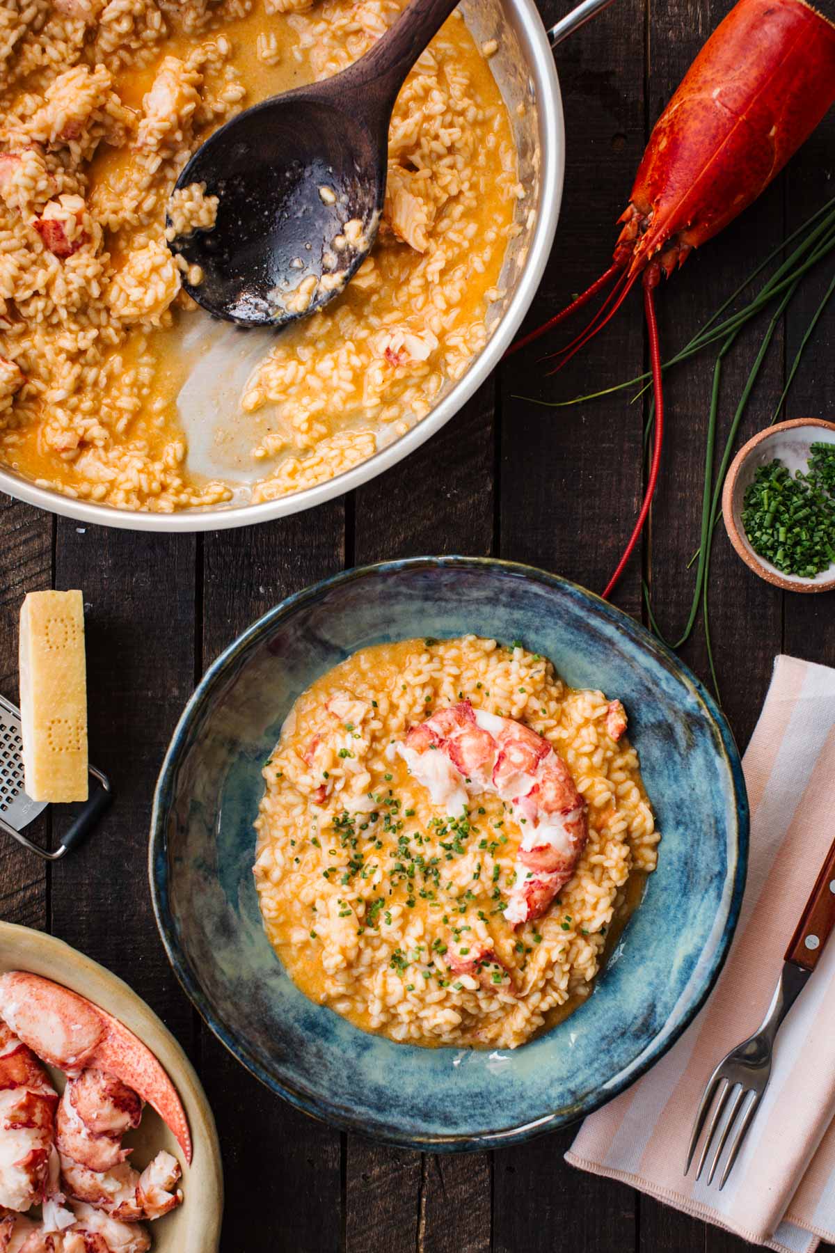 Overhead shot of lobster risotto in a steel pan with a wooden spoon, next to a bowl of finished risotto topped with a lobster tail half and chives, a whole cooked lobster, parmesan block and grater, and a small bowl of chopped chives on a dark wood table.
