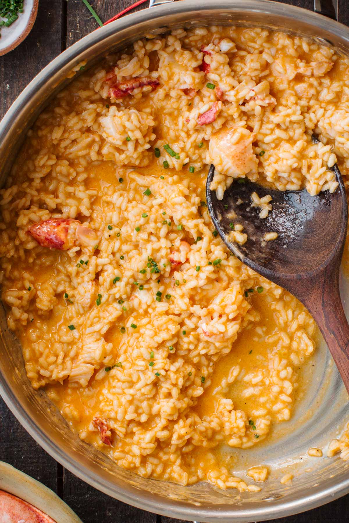 Close-up overhead view of creamy lobster risotto in a steel pan, studded with chunks of lobster meat and flecked with chopped chives, a wooden spoon resting in the pan.