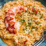 Close-up of a bowl of lobster risotto garnished with chives, with a whole lobster tail half draped across the top and a fork resting on the edge of the bowl.