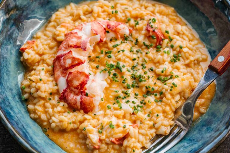 Close-up of a bowl of lobster risotto garnished with chives, with a whole lobster tail half draped across the top and a fork resting on the edge of the bowl.