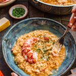 A hand holds a bowl of lobster risotto and digs in with a fork, with the pan of risotto, a bowl of chives, parmesan, and lobster shells arranged on the table nearby.