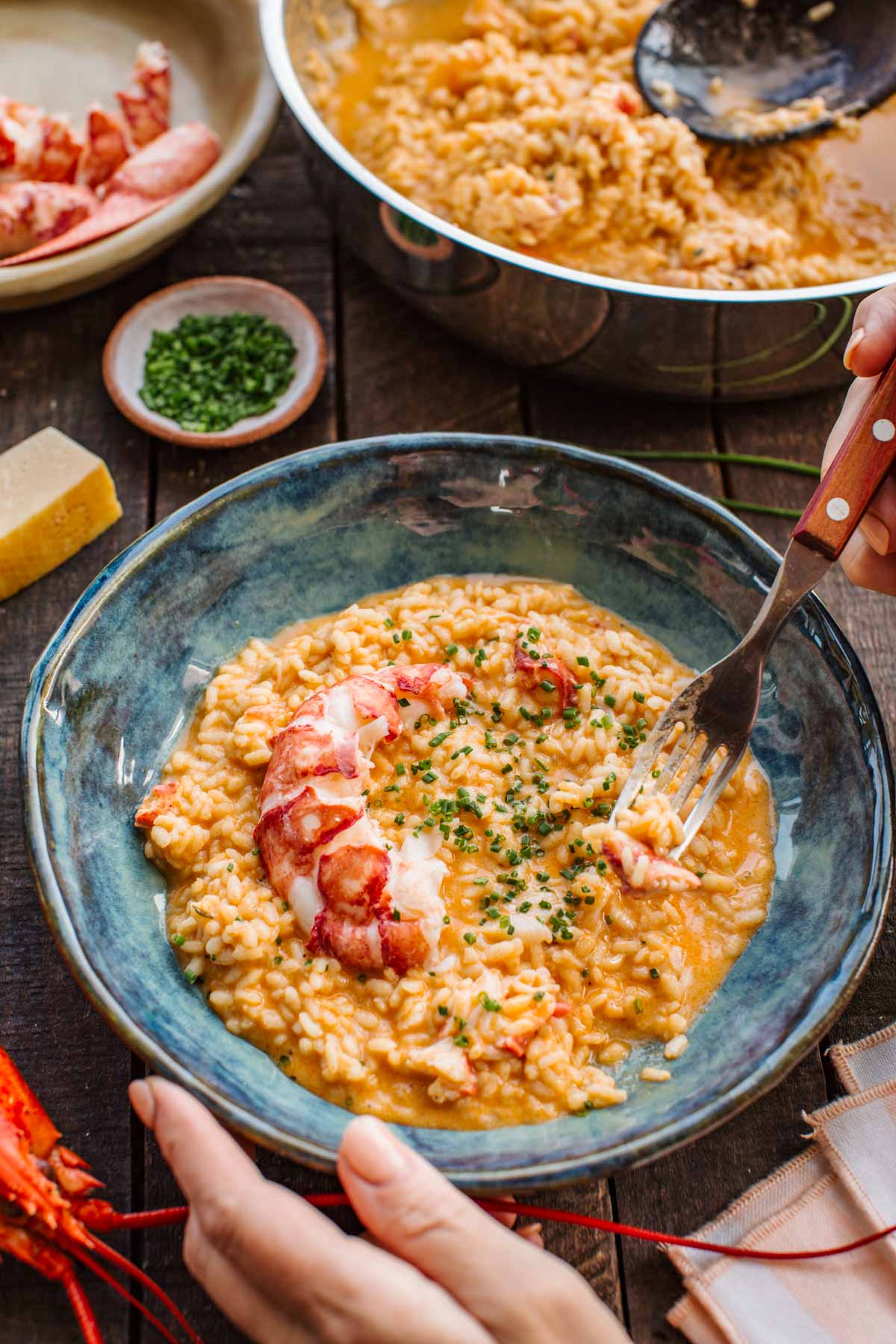 A hand holds a bowl of lobster risotto and digs in with a fork, with the pan of risotto, a bowl of chives, parmesan, and lobster shells arranged on the table nearby.