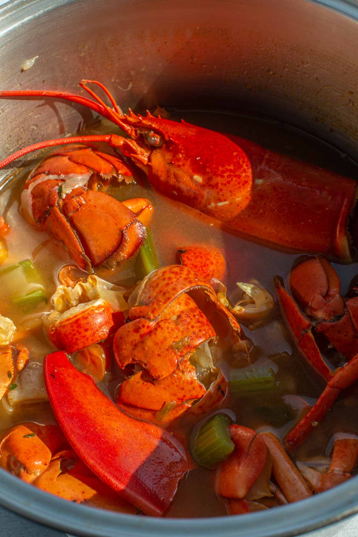 Lobster shells, celery, and carrot pieces simmering in a stainless steel pot filled with orange-hued stock.