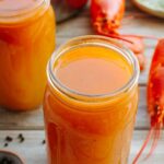 Close-up of mason jars filled with strained lobster stock, with a lobster shell and stockpot visible in the background.