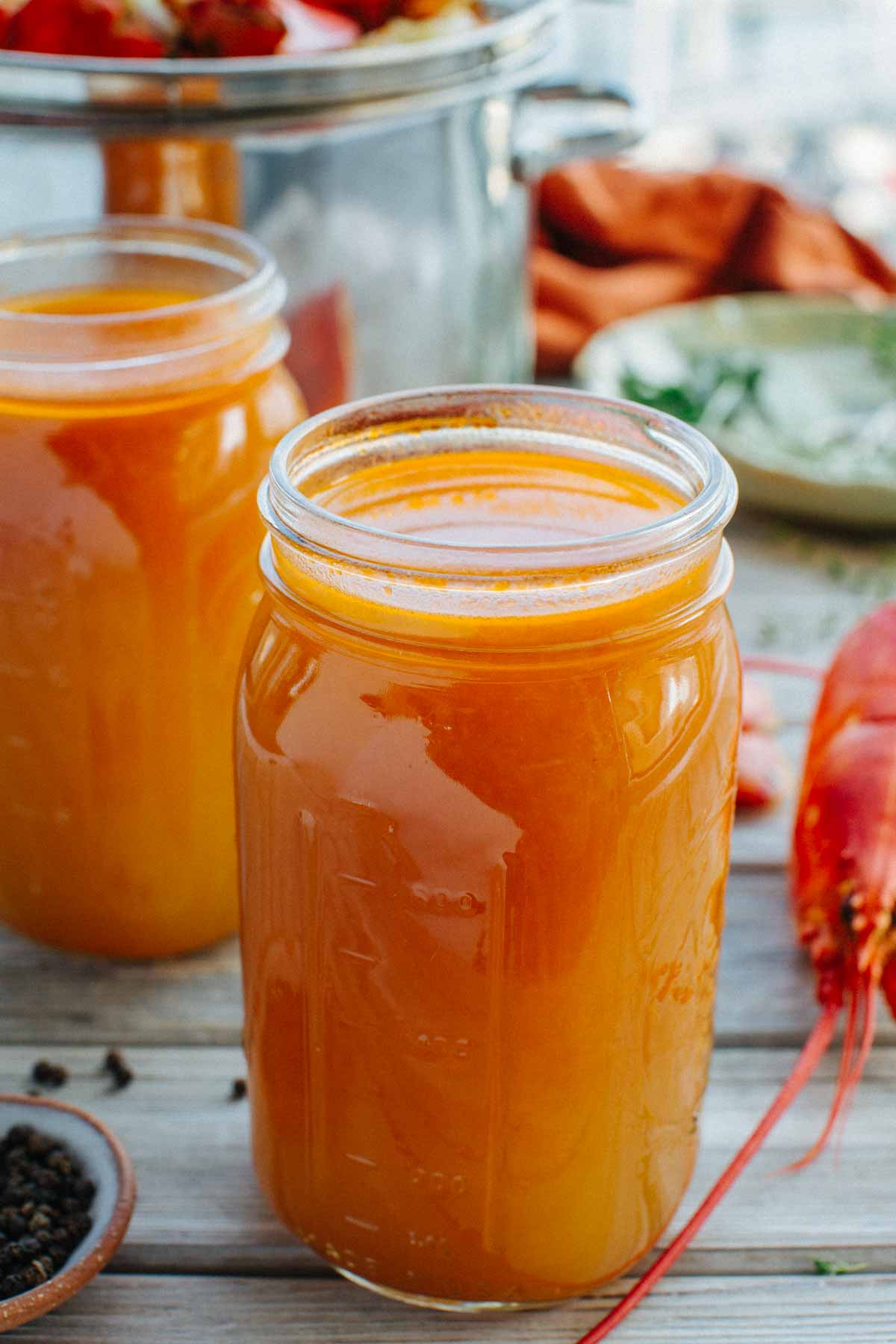 Close-up of mason jars filled with strained lobster stock, with a lobster shell and stockpot visible in the background.