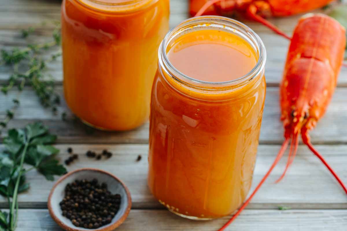 Two jars of amber lobster stock on a wooden surface, with fresh herbs, black peppercorns, and a lobster shell alongside.
