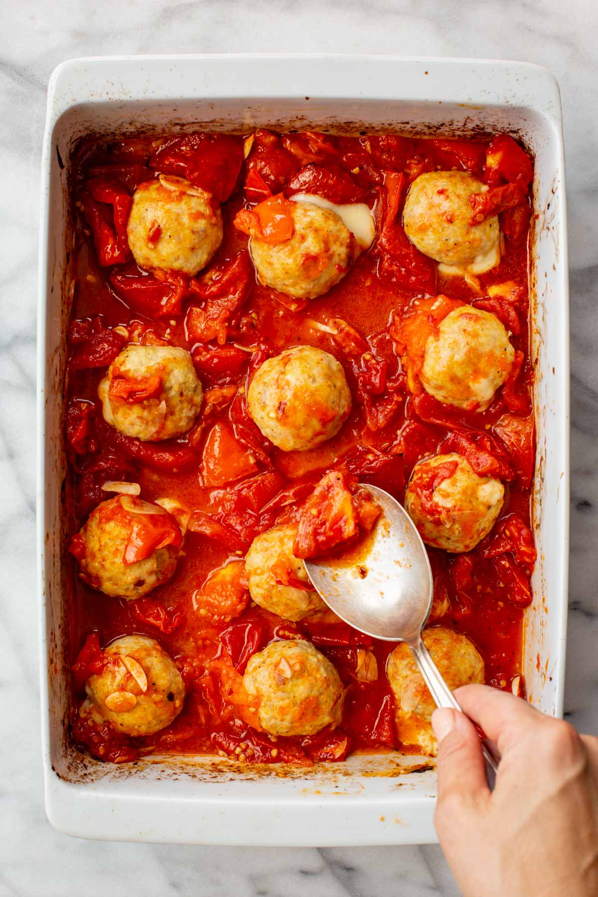 Baked chicken meatballs being spooned with roasted tomato sauce.