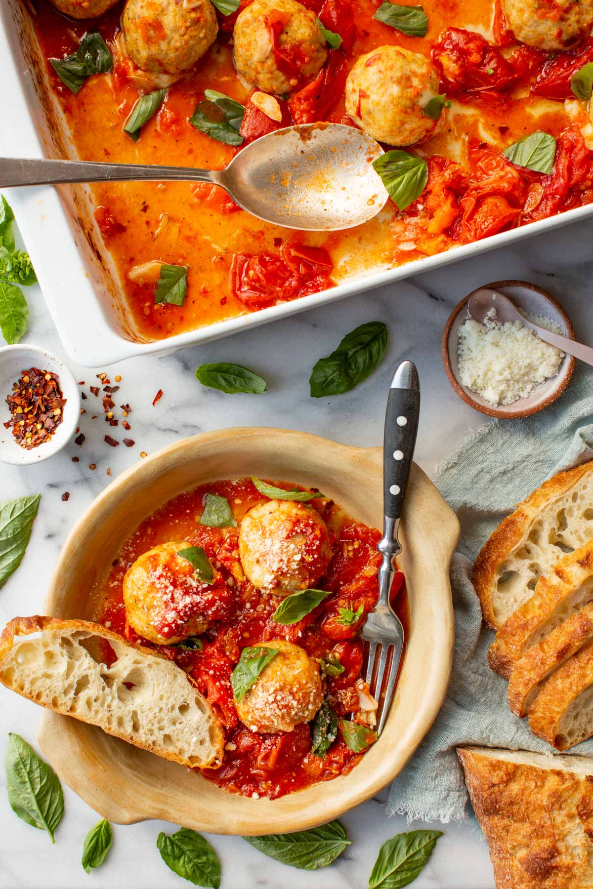 A plate of stuffed chicken meatballs with roasted tomato sauce next to a baking dish filled with meatballs, sliced bread, salt and basil leaves.