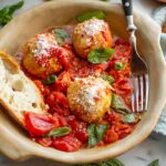 Close-up of a ceramic plate filled with chicken meatballs in roasted tomato sauce, topped with grated pecorino, next to a fork and a slice of bread.