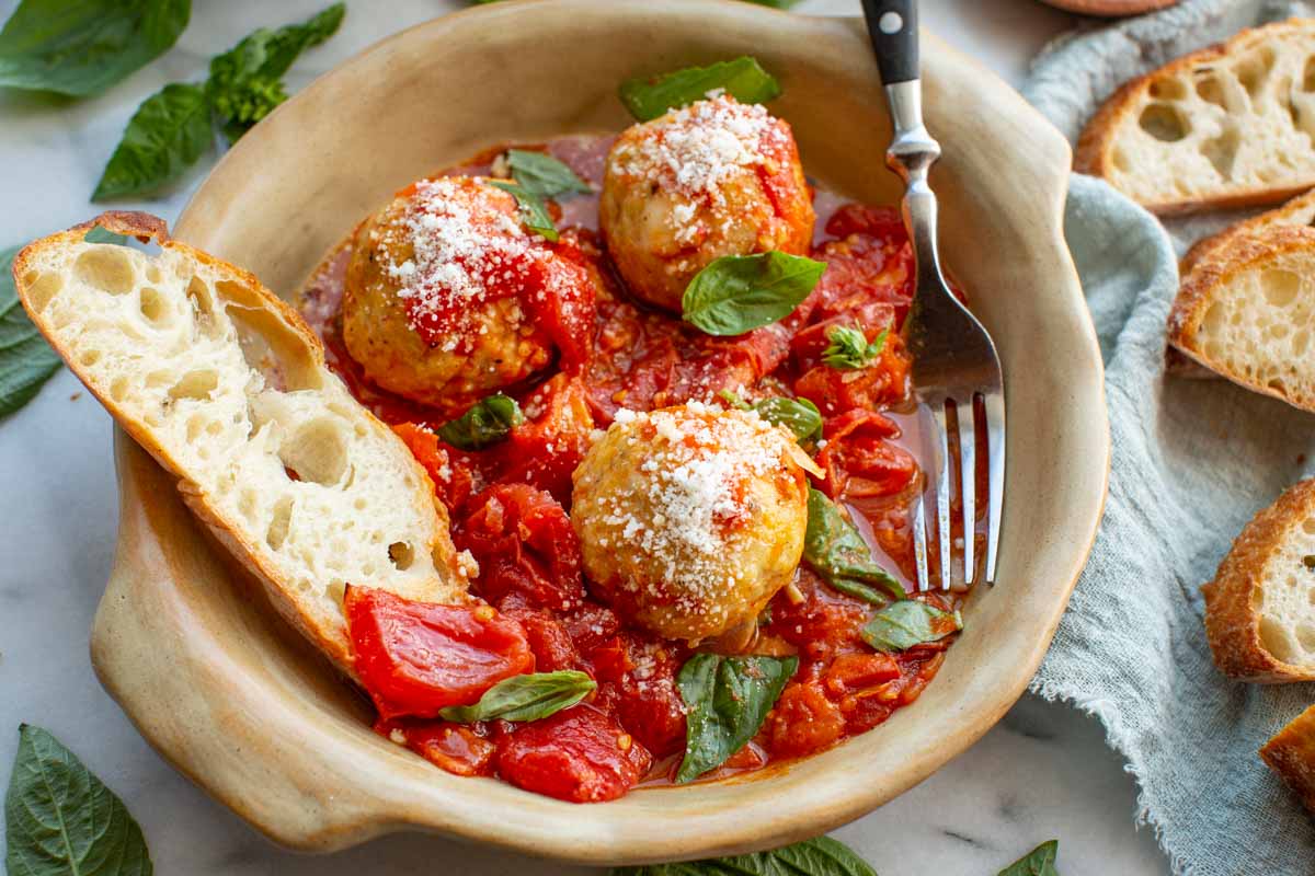 Close-up of a ceramic plate filled with chicken meatballs in roasted tomato sauce, topped with grated pecorino, next to a fork and a slice of bread.