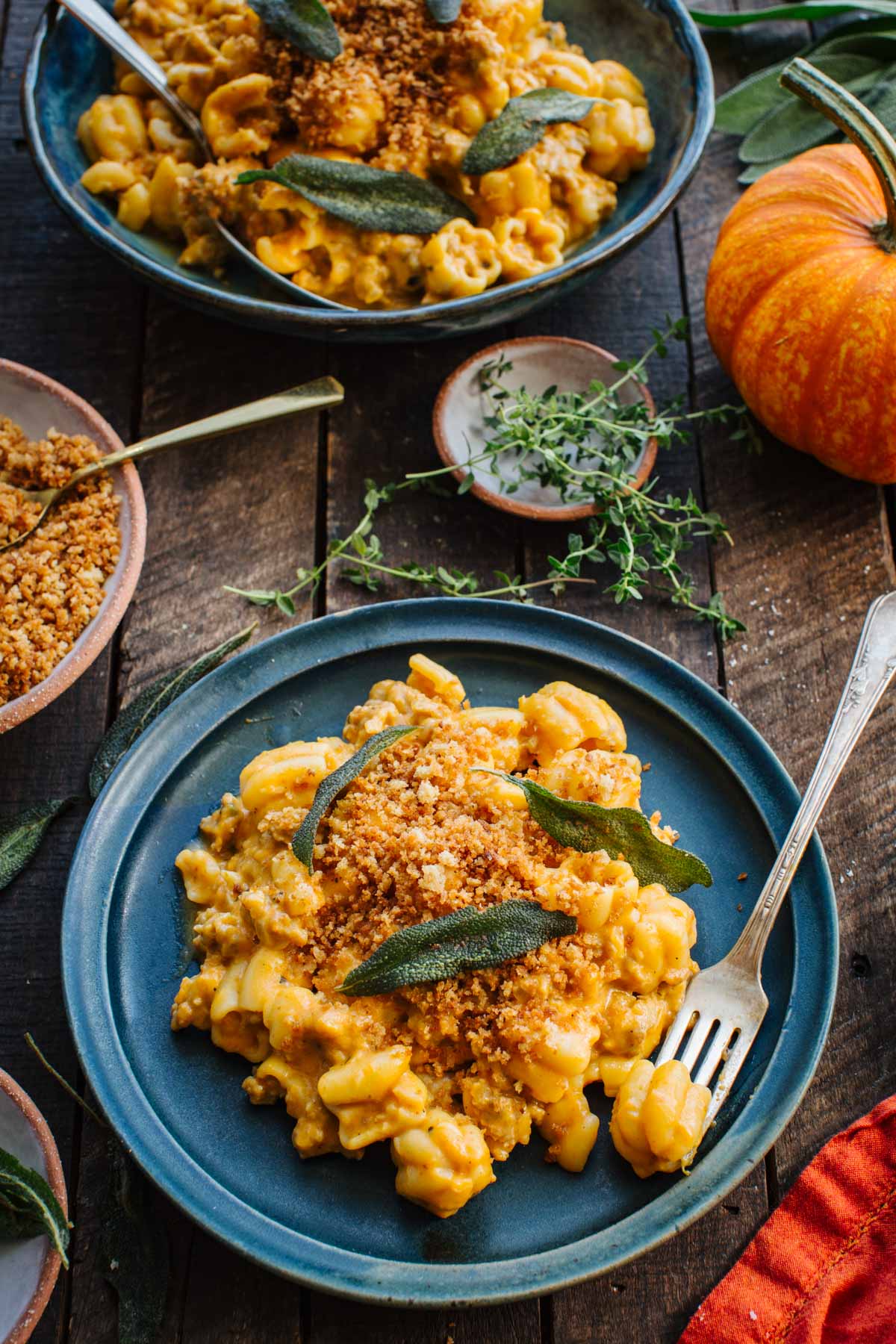 Overhead shot of pumpkin sausage pasta served on a blue plate topped with toasted breadcrumbs and fried sage leaves, with a fork, fresh thyme, a mini pumpkin, and a second bowl of pasta in the background.