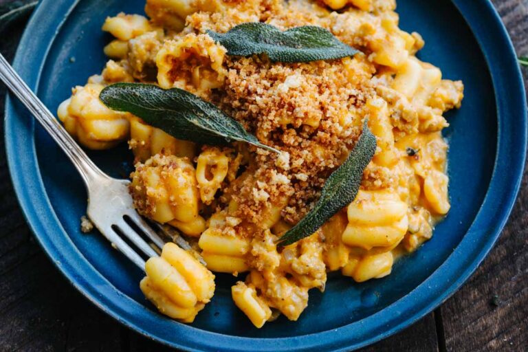 Close-up of pumpkin sausage pasta on a blue plate, garnished with crispy fried sage leaves and a golden breadcrumb topping, fork resting alongside.