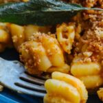 Extreme close-up of a forkful of creamy pumpkin pasta lifted from the plate, showing the pasta's ridged shape coated in sauce with toasted breadcrumbs and a fried sage leaf in the background.