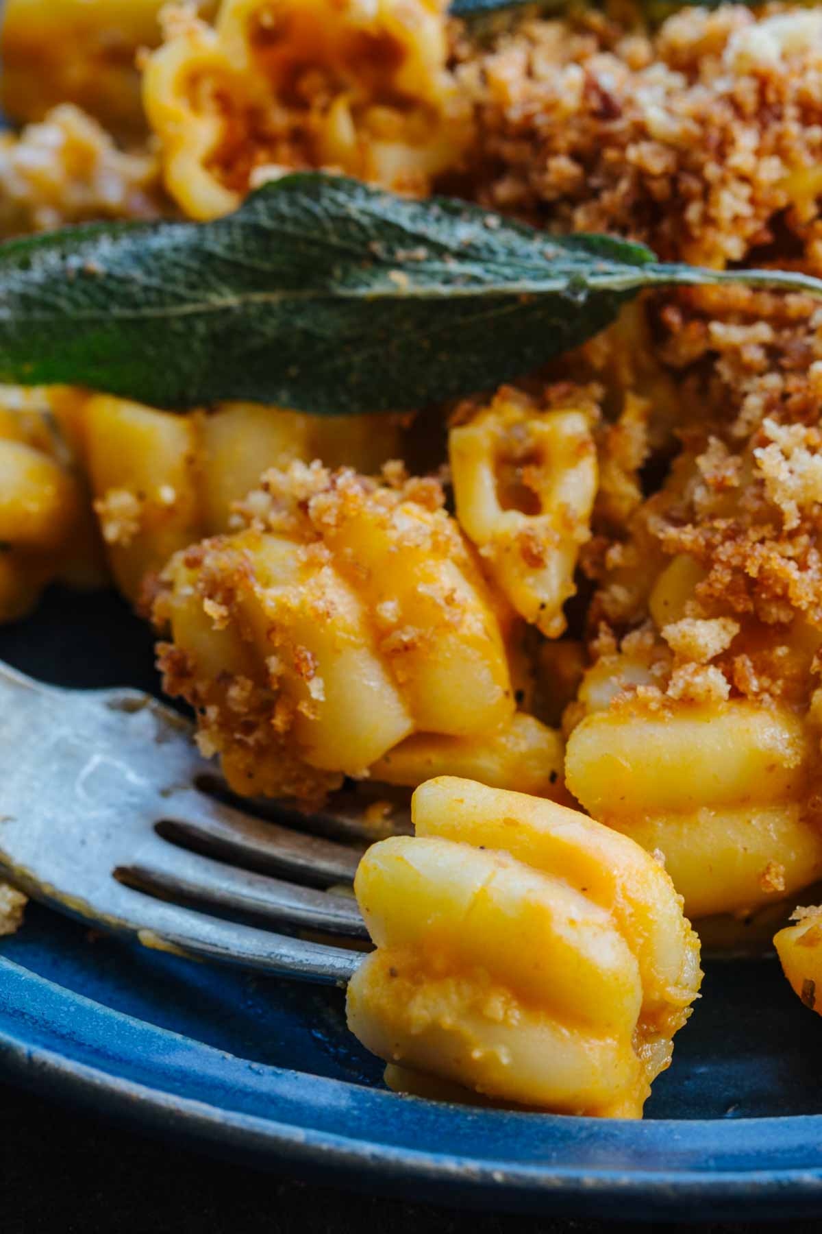 Extreme close-up of a forkful of creamy pumpkin pasta lifted from the plate, showing the pasta's ridged shape coated in sauce with toasted breadcrumbs and a fried sage leaf in the background.