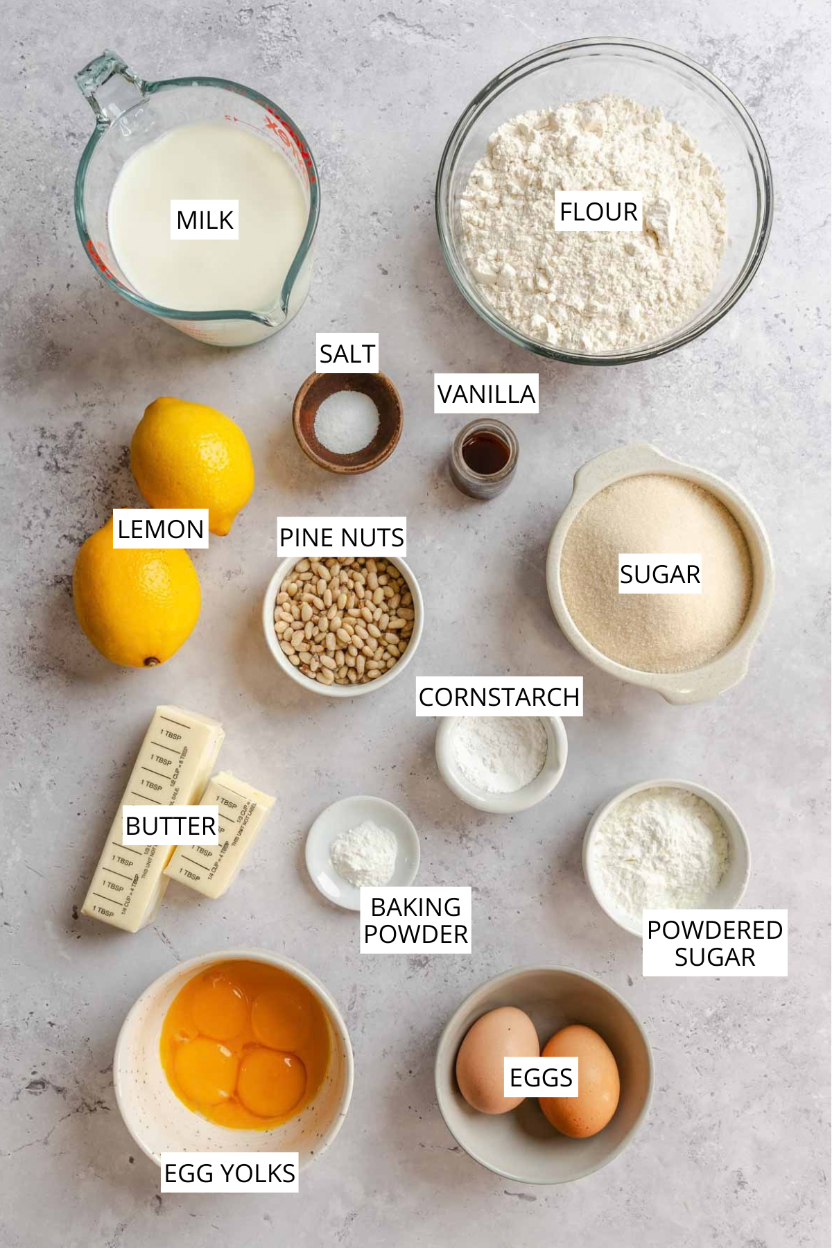 Overhead view of the labeled ingredients for torta della nonna: milk, flour, salt, vanilla, lemons, pine nuts, sugar, cornstarch, butter, baking powder, powdered sugar, egg yolks, and whole eggs.