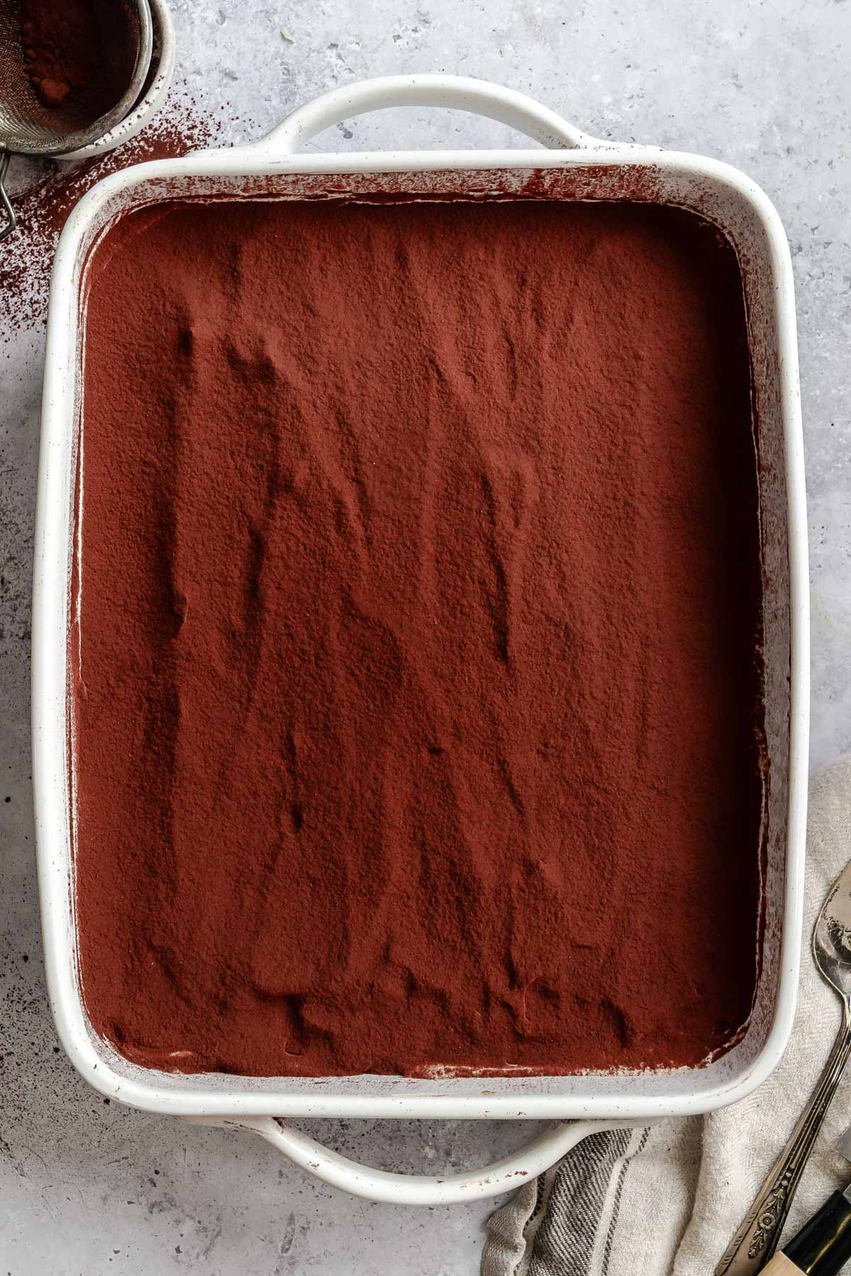 Overhead view of a white baking dish filled with tiramisu, the top evenly covered in a thick layer of dark cocoa powder.