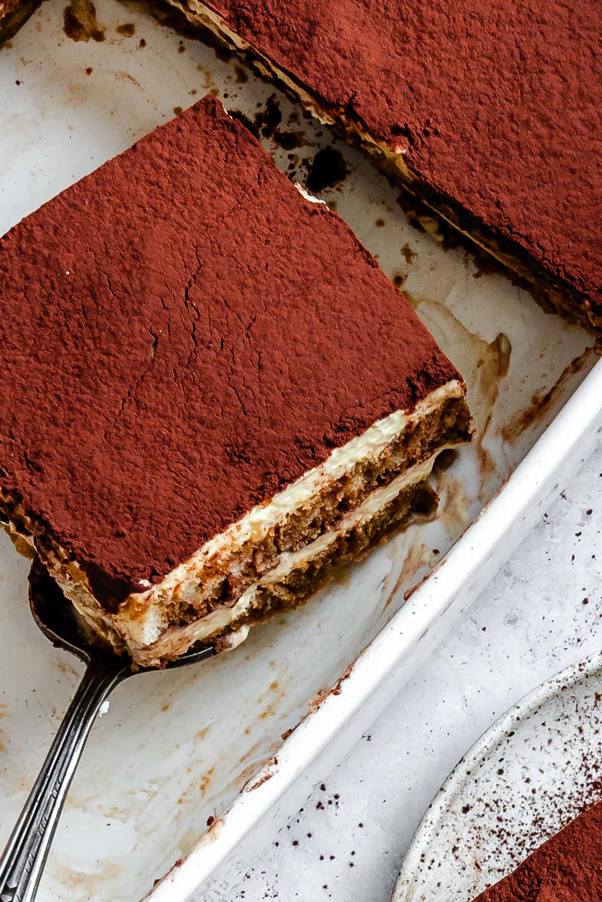 Close-up of a square slice of tiramisu on a serving spatula, showing cocoa on top and two layers of soaked ladyfingers separated by creamy filling.