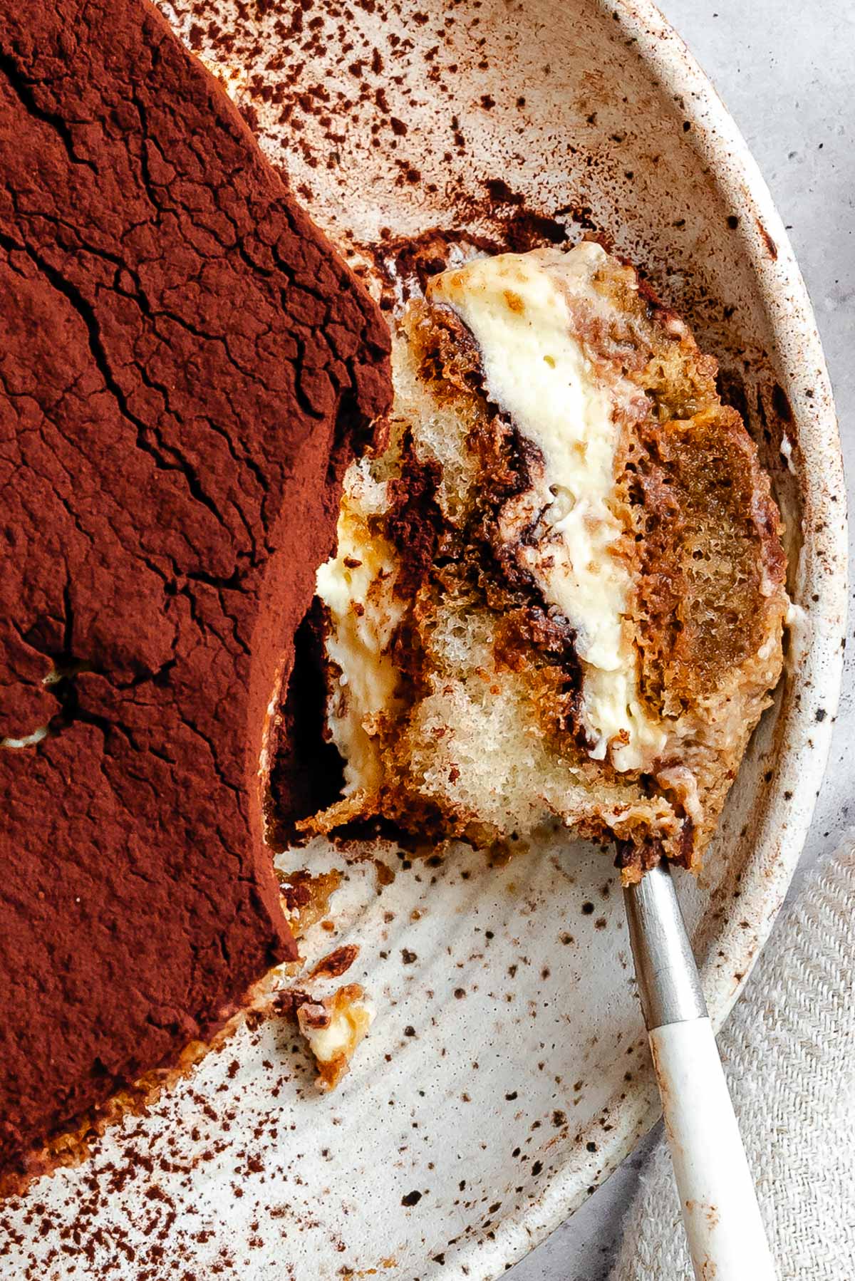 Overhead close-up of a cocoa-covered tiramisu slice on a speckled plate with a forkful pulled away, revealing soft ladyfingers and layers of mascarpone cream.