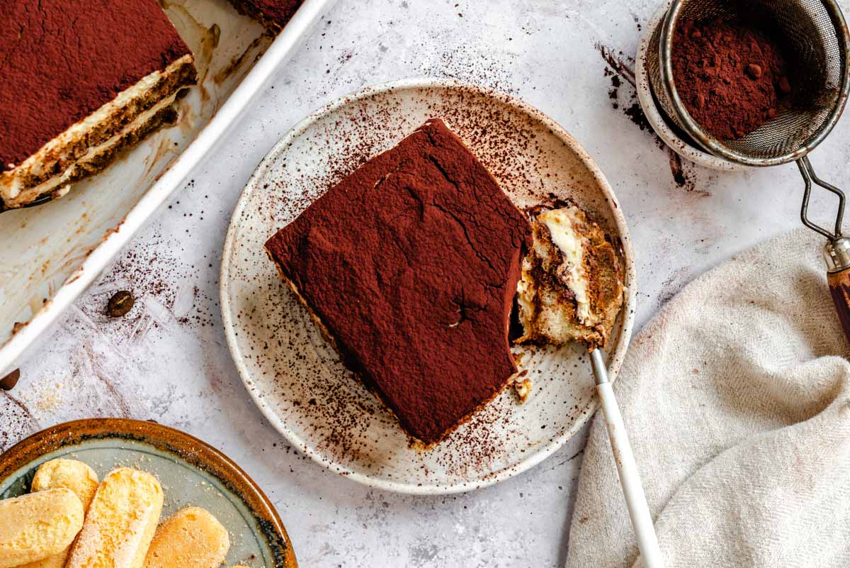 Overhead view of a tiramisu slice on a speckled ceramic plate with a fork bite taken out, with the baking dish, a small sieve of cocoa, a linen napkin, and a plate of ladyfingers around it.
