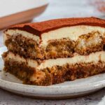Side view of a tiramisu slice on a speckled plate, showing a cocoa-dusted top, a thick mascarpone cream layer, and two layers of coffee-soaked ladyfingers.