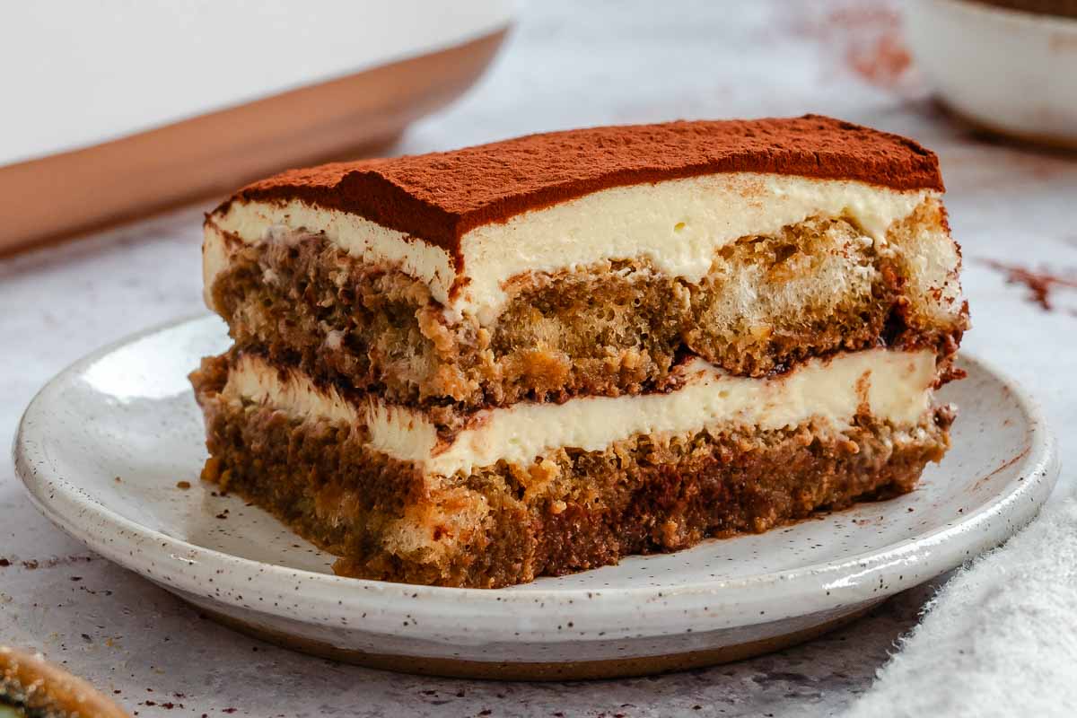 Side view of a tiramisu slice on a speckled plate, showing a cocoa-dusted top, a thick mascarpone cream layer, and two layers of coffee-soaked ladyfingers.