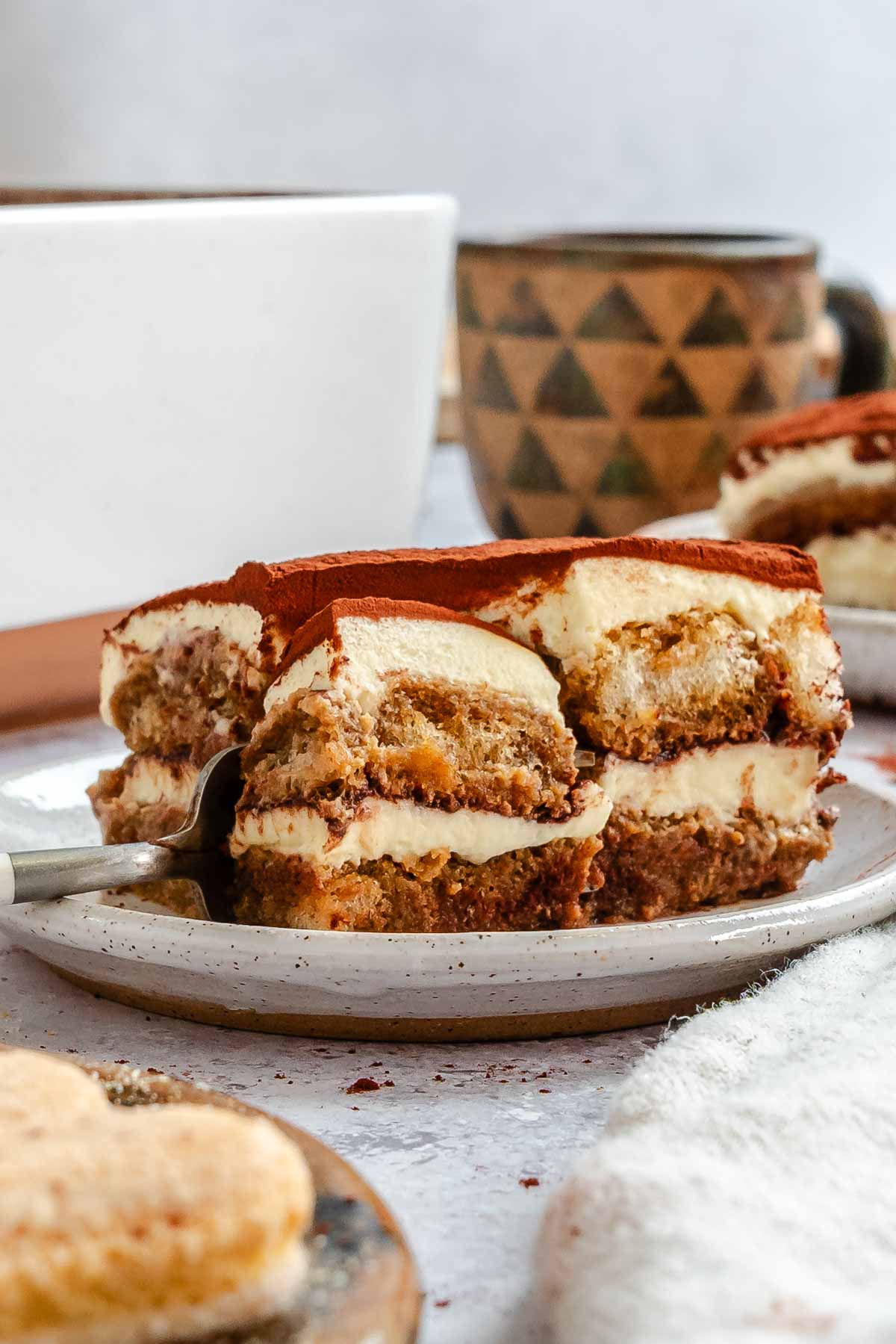 Side view of a tiramisu slice on a speckled plate with a fork cutting into it, with a white baking dish and a patterned ceramic mug in the background.