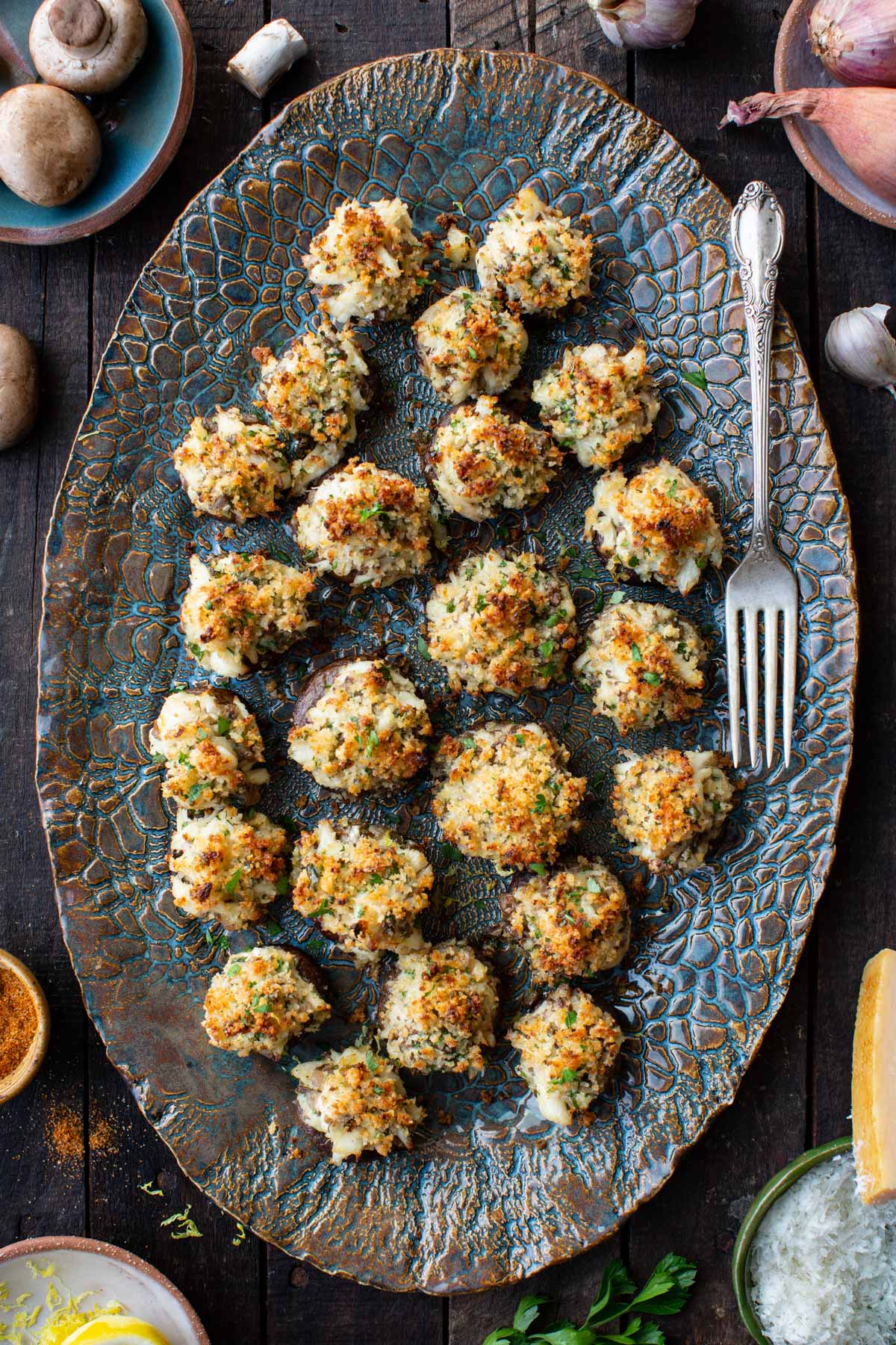 Overhead view of crab stuffed mushrooms on a blue textured oval platter with a silver fork, surrounded by raw mushrooms, shallots, garlic, parmesan, lemon, and parsley.