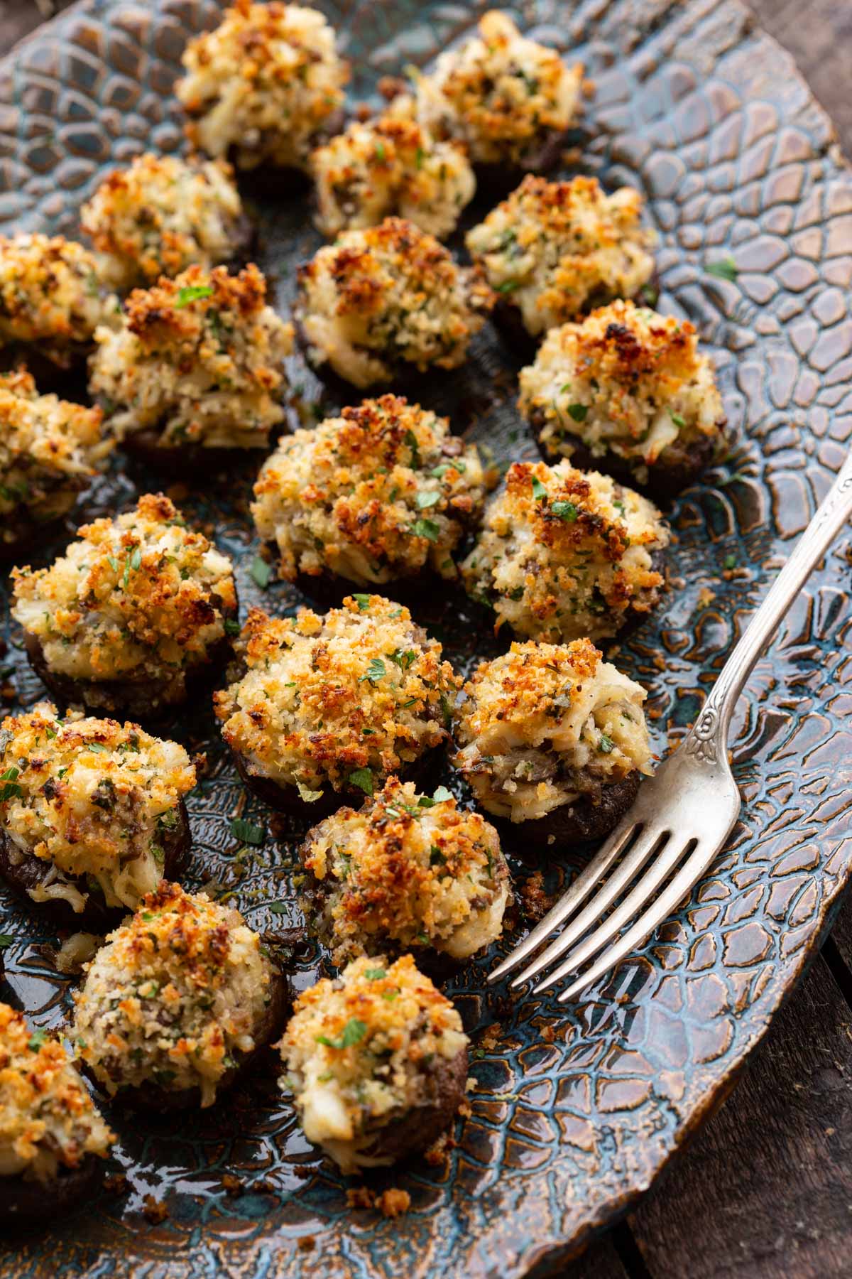Angled view of crab stuffed mushroom appetizers with browned breadcrumb topping on a blue patterned serving platter beside a silver fork.