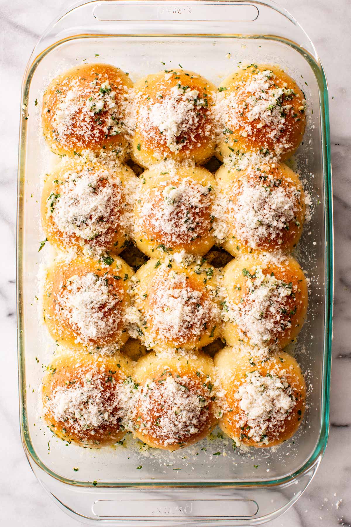 Overhead view of twelve golden garlic parmesan dinner rolls in a glass baking dish, topped with grated parmesan, minced garlic, and parsley.