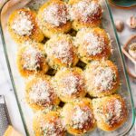 Pan of garlic parmesan dinner rolls surrounded by a wedge of parmesan on a grater, a dish of butter, garlic bulbs, and chopped parsley.