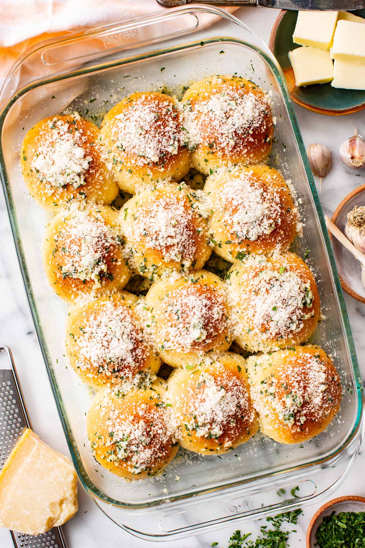 Pan of garlic parmesan dinner rolls surrounded by a wedge of parmesan on a grater, a dish of butter, garlic bulbs, and chopped parsley.