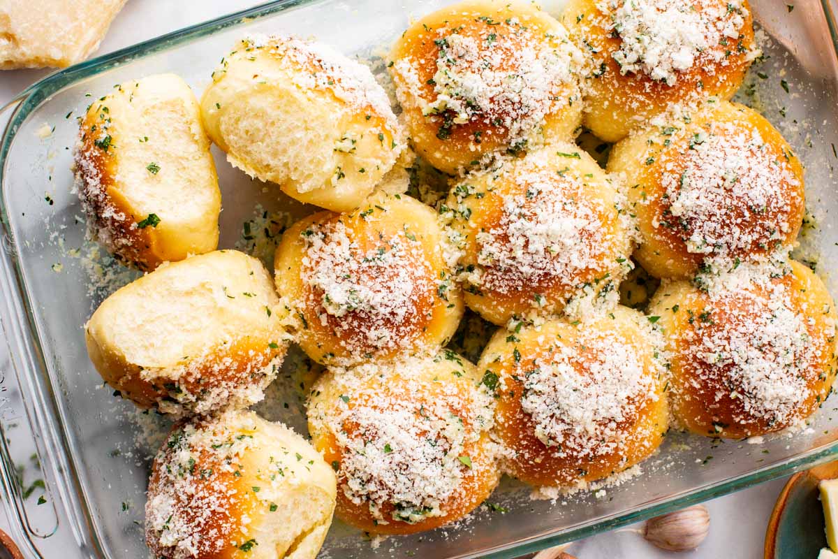 Baked garlic parmesan rolls in a glass dish, with a few pulled apart to show the soft, fluffy interior.