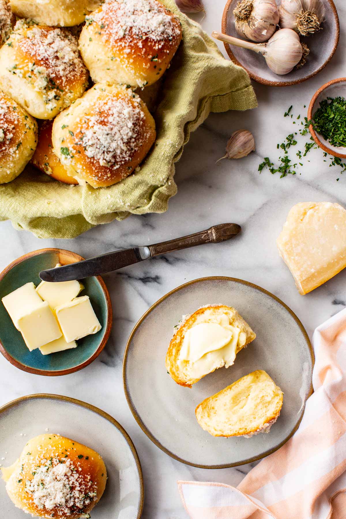 Garlic parmesan rolls in a green linen-lined basket beside a dish of butter, a roll split open and buttered on a plate, garlic, parsley, and a parmesan wedge.