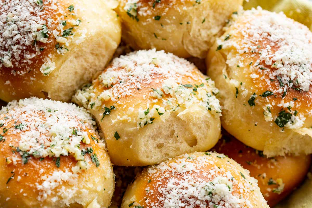 Close-up of soft garlic parmesan rolls with a golden crust, parmesan and parsley topping, and a pulled-apart roll showing the airy crumb.