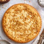 Overhead view of a whole golden Italian custard tart with a fluted crust, its top covered in toasted pine nuts, surrounded by a bowl of pine nuts, a bowl of powdered sugar, a small sieve, vintage spoons, and a linen napkin.