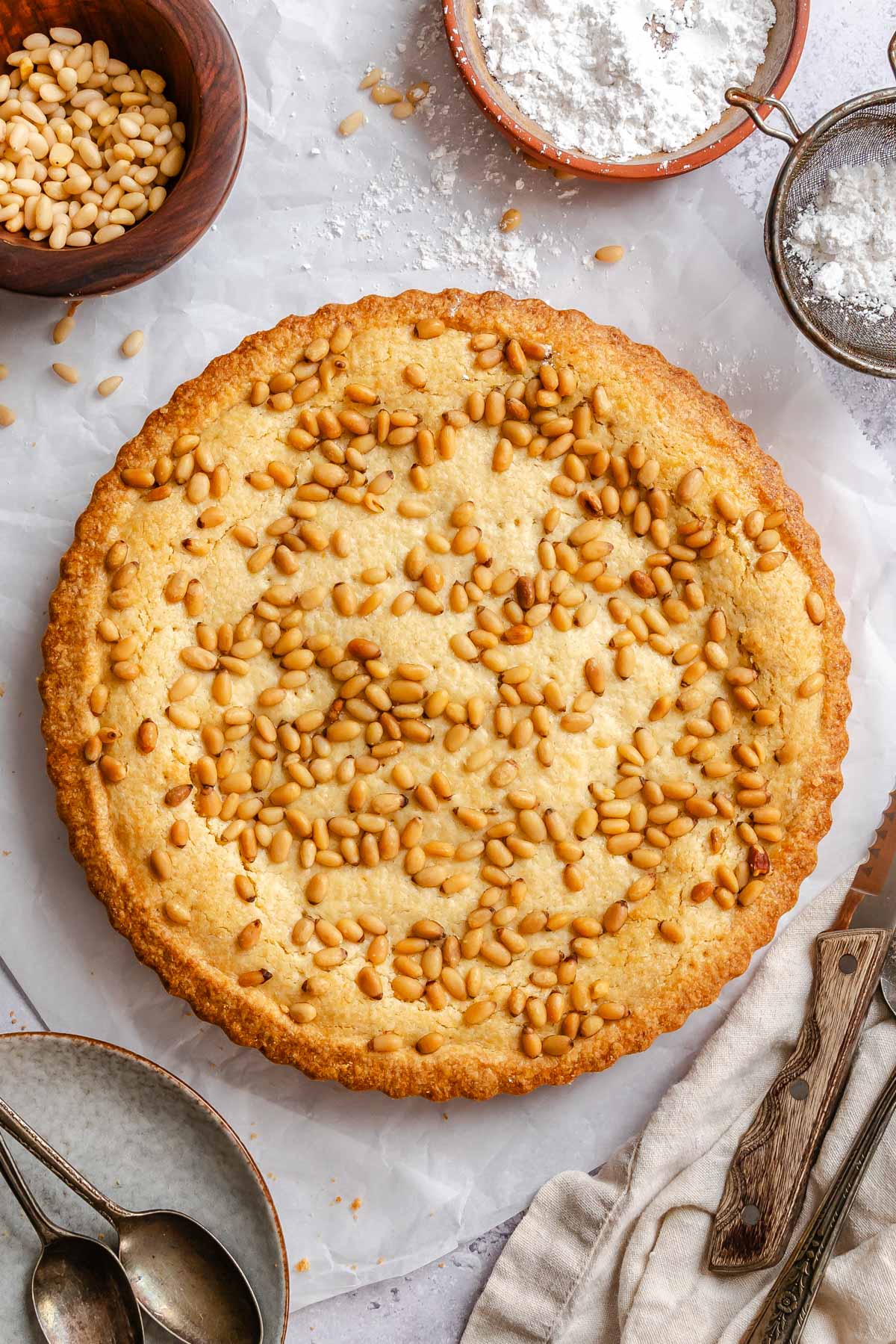 Overhead view of a whole golden Italian custard tart with a fluted crust, its top covered in toasted pine nuts, surrounded by a bowl of pine nuts, a bowl of powdered sugar, a small sieve, vintage spoons, and a linen napkin.