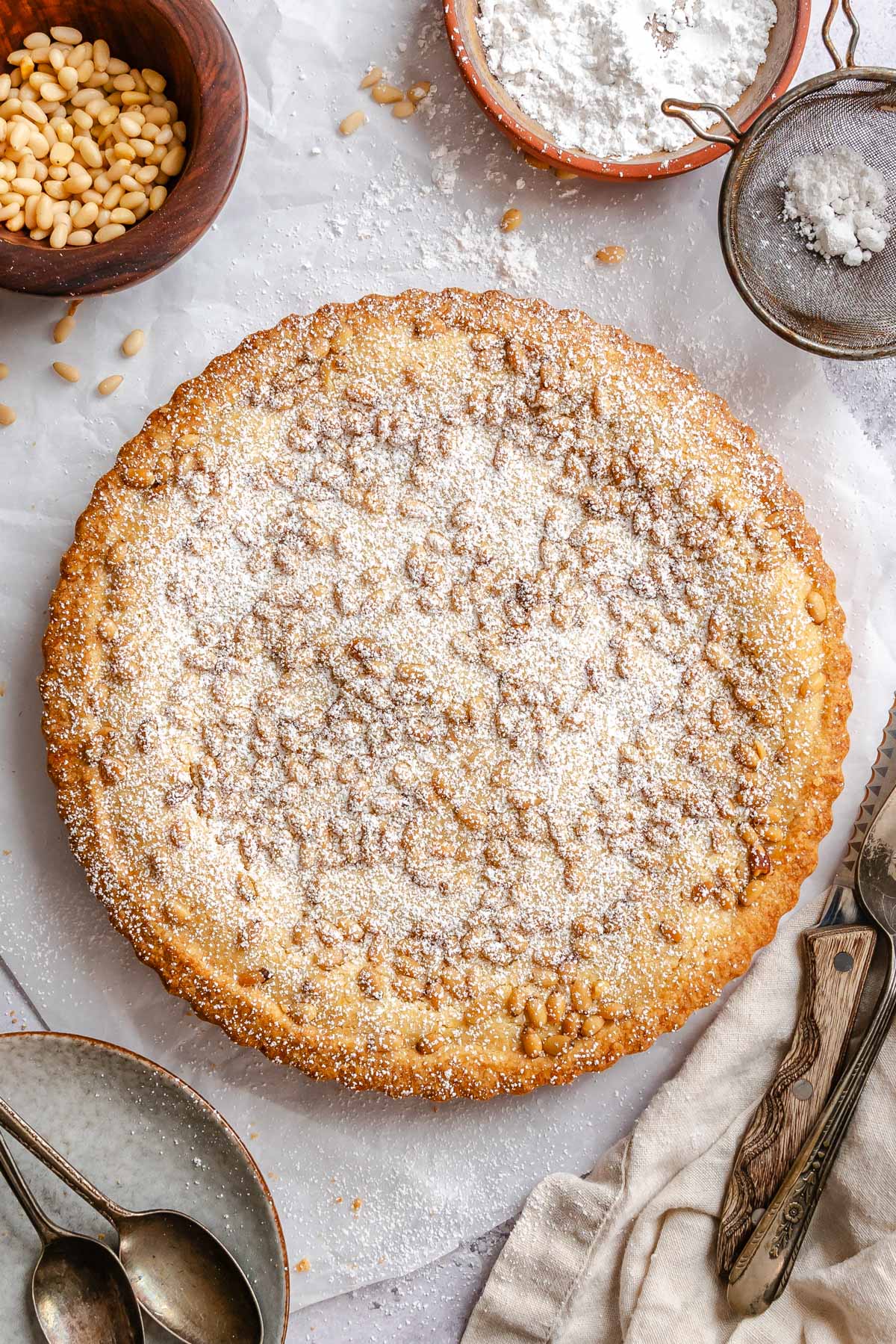 Overhead view of a whole torta della nonna with a golden fluted crust, its pine nut topping lightly dusted with powdered sugar.