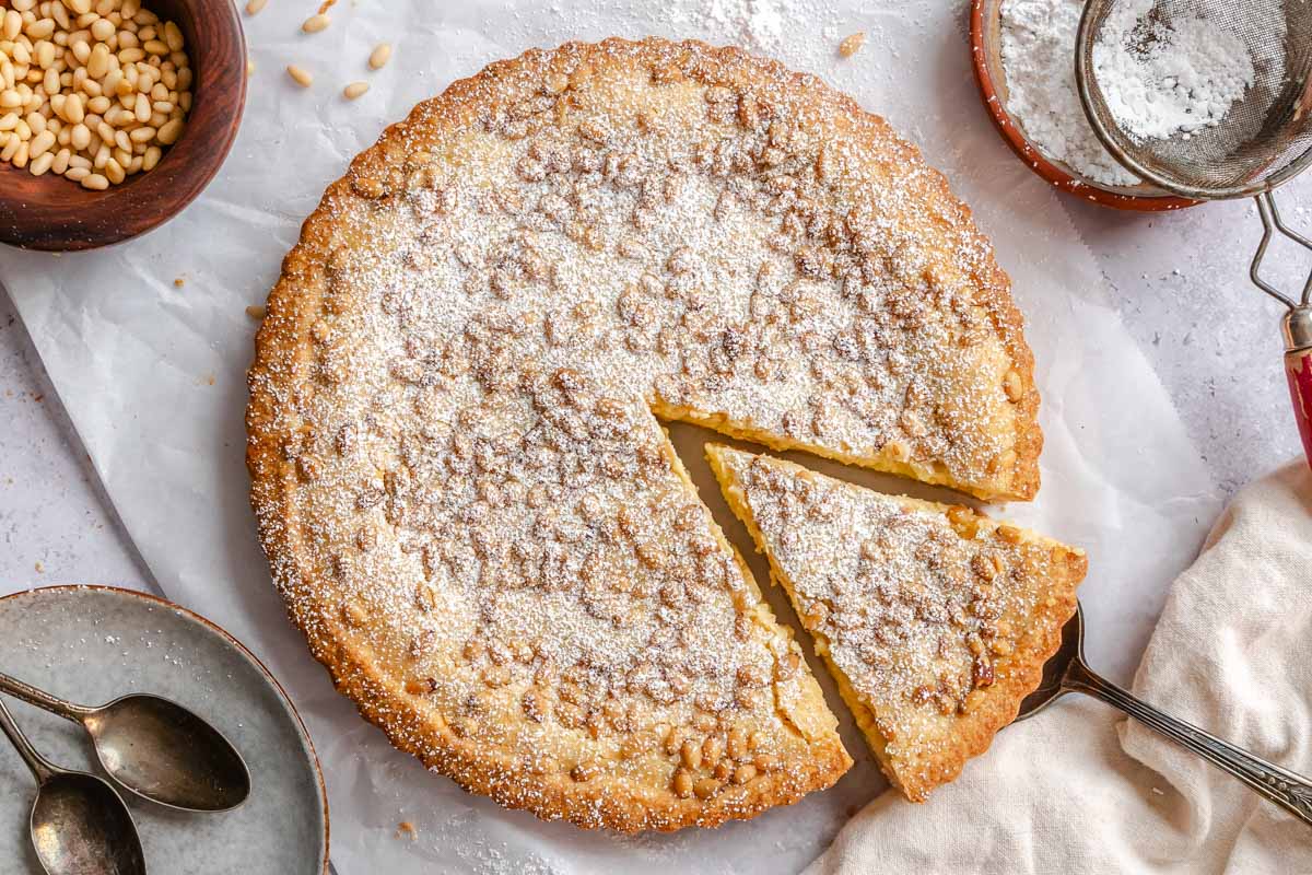 Overhead view of a powdered sugar-dusted pine nut custard tart with one slice cut and lifted slightly out on a vintage server.